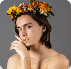 jovem de pele clara, cabelos escuros e compridos, posando para a foto com uma das mãos apoiadas na face, e usando uma coroa de flores na cabeça